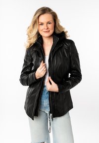 Blonde vrouw die een zwart leren jack over een witte top en verlichte blauwe jeans draagt, met beide handen de rits van het jack vasthoudend.