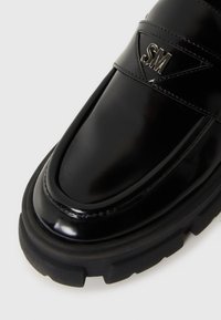 Pantofii din piele lacuită neagră au vârful rotund, talpă groasă din cauciuc și un accent metalic proeminent cu logo. Prezintă o textură netedă și un design elegant.