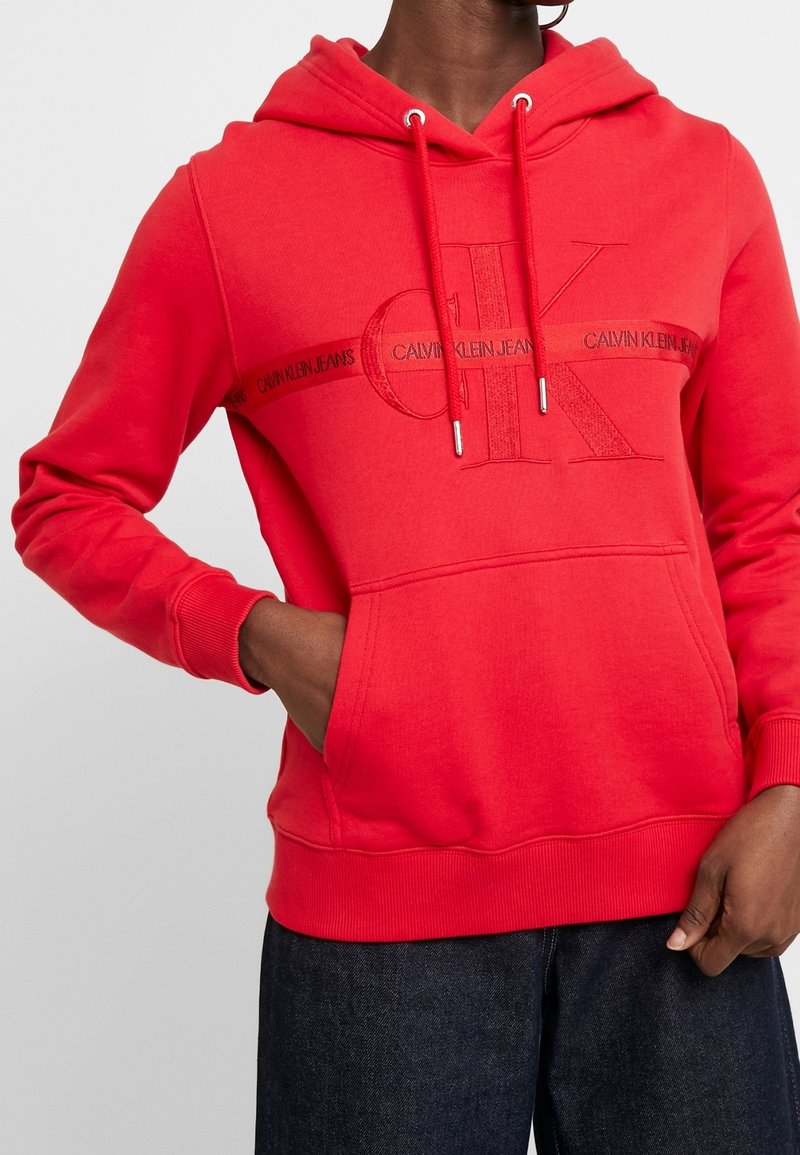 Personne portant un sweat à capuche Calvin Klein rouge vif avec poche avant, cordons de serrage et grand logo CK embossé sur la poitrine.