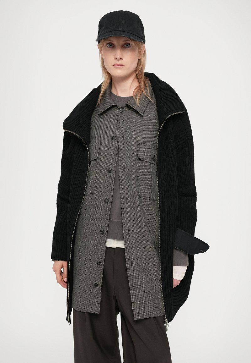 Abrigo negro de punto oversize con cierre frontal, superpuesto a una camisa de cuadros gris con botones. Presenta una gorra negra y pantalones oscuros de corte holgado.