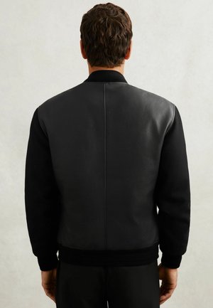 Man die met zijn rug naar de camera staat, draagt een zwarte jas met een gestructureerd leren achterpaneel en gladde stoffen mouwen tegen een effen lichte achtergrond.