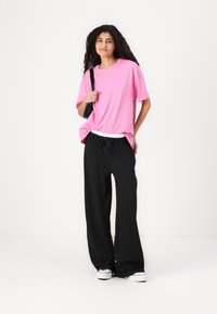 T-shirt en coton rose associé à un pantalon large à rayures noires et une ceinture blanche. Le mannequin tient un sac à bandoulière noir et porte des baskets blanches.