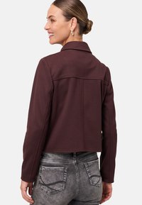 Veste courte bordeaux avec un col classique, coupe ajustée et texture lisse, associée à un jean léger avec poches arrière.