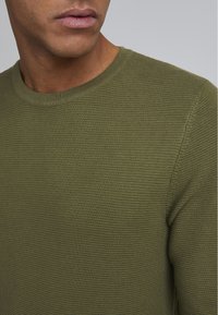 Pulover tricotat de culoare verde măslinie, cu un neckline rotund și țesătură texturată. Vedere detaliată care subliniază cusătura și textura fibrelor.