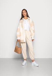 Romige en tan geruite fleecejack, wit t-shirt, beige joggers, witte sneakers en een bruine handtas met een gekreukt handvat.