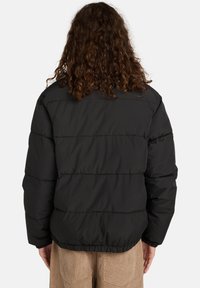 Person med krøllet hår iført en sort pufferjakke og beige corduroypants, vist fra ryggen mod en ensfarvet baggrund.