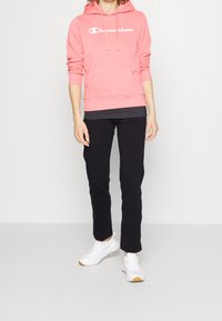 Rosa hoodie med vitt logotyp, svarta byxor och vita sneakers med beige accenter. Hoodien har en ficka fram och justerbar dragsko.