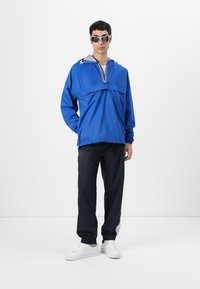Blå lett hettejakke med grå hette og flerfarget glidelås, kombinert med sorte bukser og hvite joggesko.