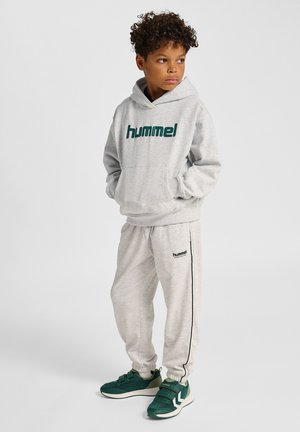 Jasnoszara bluza z kapturem z logo "hummel", do tego dopasowane szare spodnie dresowe z elastycznymi mankietami oraz zielone sneakersy z paskiem. Minimalistyczny design.