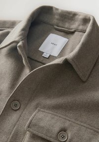 Ruskea villasekoitepaita, jossa on viistot kaulukset, kaksi napitettavaa rintataskua ja harmaat napit. Tunnuksessa lukee "NEXT," koko M. Pehmeä rakenne.