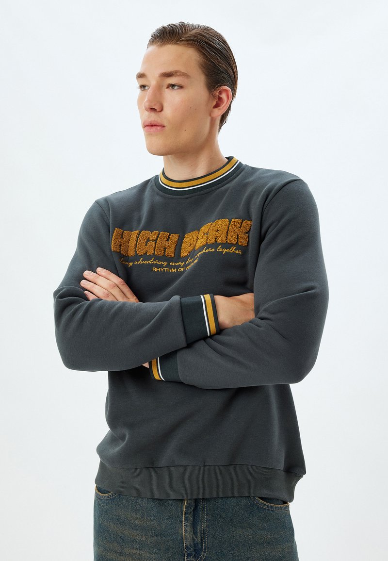Sudadera gris oscuro con cuello redondo acanalado, con rayas amarillas y blancas. En la parte frontal tiene letras en relieve "HIGH PEAK" en negrita. Tejido suave.