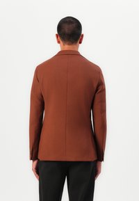 Sacou brun bine croit, cu o textură netedă și umeri structurați. Prezentă o deschidere unică și manșete cu nasturi, asortat cu pantaloni închiși la culoare.