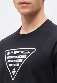Νεαρός ενήλικας που φοράει μαύρο T-shirt με λευκό τρίγωνο λογότυπο που γράφει "PFG Performance Fishing Gear" στο στήθος, περικομμένο στον λαιμό και τους ώμους.