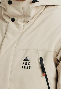 Veste imperméable beige avec un col haut, des boutons-pression noirs et une poche zippée. Présente le logo "PRO TEST" en noir sur la zone de la poitrine.