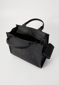 Zwarte leren DKNY-handtas met reliëflogo, twee handvatten, open bovenkant met zichtbare gouden rits binnenin, en een bevestigd klein zakje aan de zijkant.