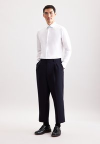 Camicia bianca con bottoni e maniche lunghe, abbinata a pantaloni a pieghe blu navy e scarpe di pelle nera. Design semplice e sartoriale con dettagli minimi.