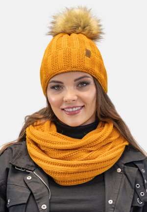 Orange knitted hat with a faux fur pom-pom and a textured design, paired with a matching knitted scarf in the same color. Soft, warm materials.