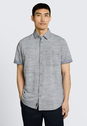 Mann trägt ein kurzärmeliges, graues Hemd mit Knöpfen und einer Brusttasche, steht mit einer Hand in der Tasche.