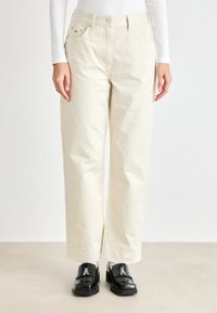 Beige vide benbukser med høy midje, laget av bomullsstoff. Har frontlommer og knapper i sølvfarge. Sorte loafers brukes.