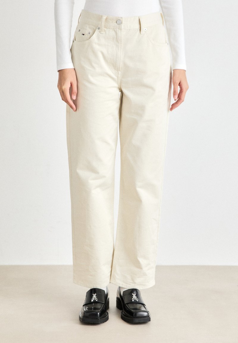 Beige vide benbukser med høy midje, laget av bomullsstoff. Har frontlommer og knapper i sølvfarge. Sorte loafers brukes.