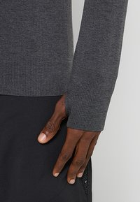 Długi rękaw ciemnoszarej bluzki o prążkowanej fakturze, z otworem na kciuk przy mankiecie. Ręka modelki spoczywa na czarnym, teksturowanym dolnym elemencie garderoby.