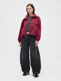 Jachetă roșie ușoară cu fermoar, guler înalt și buzunare frontale, asortată cu blugi negri cu picioare largi și cusături contrastante, și pantofi sport negri.