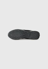 Black athletic shoe sole with a zigzag tread pattern, textured rubber material, and engraved logo detail near the heel.