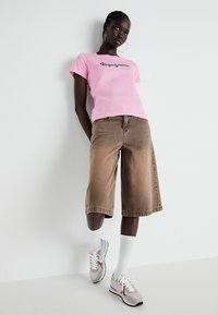 Camiseta de manga corta rosa con logotipo, pantalones cortos marrones de medio largo y zapatos deportivos grises. El modelo está de pie contra una pared blanca.