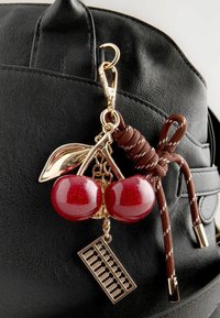 Een zwarte tas heeft een gouden kers charm met twee rode, glinsterende kersen en een decoratief blad, naast een bruine knoopdetail.