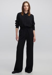 Zwarte jumpsuit met een ontwerp van een kraagshirt, lange mouwen en een elastische taille. Glad textuur, lichte glans en minimalistische knoopdetails.