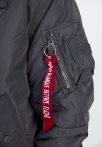Jachetă bomber din nylon gri închis, cu un buzunar cu fermoar, țesătură texturată și un etichete roșu cu textul "ÎNLEVAȚI ÎNAINTE DE ZBOR".