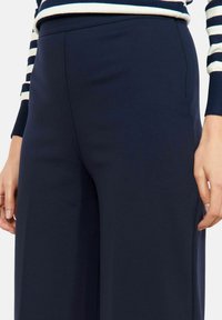 Tumesinised laia lõikega püksid, millel on sile tekstuur, kõrge vöökohaga ja küljetaskutega. Rätsepatööna lõigatud siluett parandab mugavust ja istuvust.