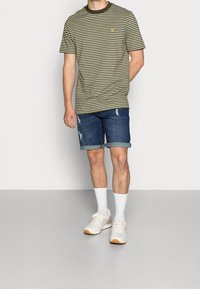 Camiseta de color verde oliva a rayas, de cuello redondo, combinada con pantalones cortos de mezclilla enrollados, de lavado claro con detalles desgastados. Calcetines blancos y zapatillas completan el look.