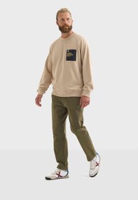 Sudadera beige con un bolsillo azul marino, pantalones verde oliva y zapatillas blancas con acentos rojos. Ajuste casual, colores sólidos y diseño simple.