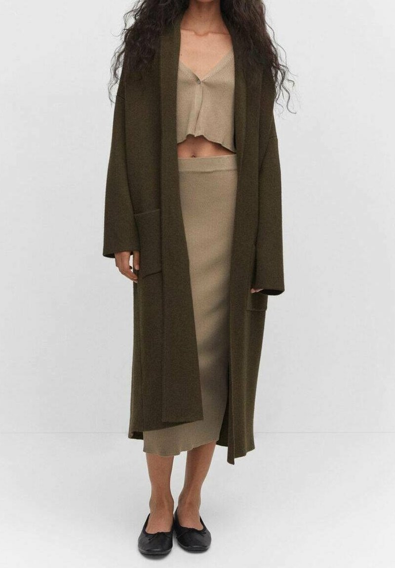 Olivenfarvet lang cardigan med lommer, lagd over en beige cropped top og nederdel, med ribbet struktur. Sorte flade sko fuldender looket.