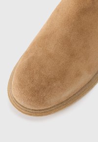 Tan ruskindsankelstøvle med rund tå, udstyret med lagdelt brun gummisål og blød struktureret overflade. Ingen synligt hardware eller detaljer.