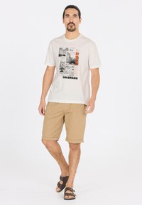 Ljust beige grafiskt t-shirt med en svart och orange tryck med bergsmotiv, tillsammans medihjullade khakishorts och bruna sandaler.