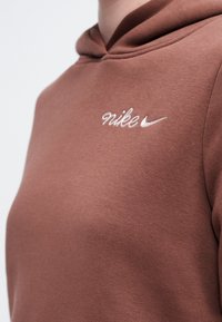 Smeđi dukserica s bijelim Nike logom izvezelim na prsima, prikazuje gornji deo trupa i područje kapuljače.