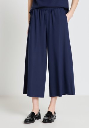 Person wearing loose navy blue cropped wide-leg pants and black leather slip-on shoes standing against a plain light background.