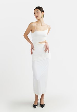 Donna con abito midi bianco arricciato senza maniche con spalline sottili, taglio in vita, orecchini statement dorati e tacchi neri a punta con fiocchi.