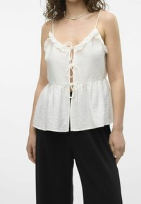 Bílý, volánkový top bez rukávů se zavázáním vpředu a rozšířeným lemem, doplněný širokými černými ribbed kalhotami.