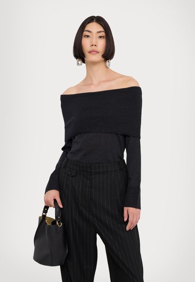 Haut en tricot noir à épaules dénudées associé à un pantalon noir à rayures. Le mannequin tient un petit sac seau noir avec des accents dorés.