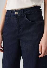 Jeans in denim blu scuro con taglio dritto, dotati di tasche frontali, un bottone metallico e dettagli di cucitura sottili. Texture liscia.