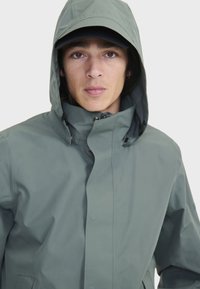 Veste imperméable vert clair avec capuche, dotée d'une fermeture à pression à l'avant et de poches latérales. Texture lisse et design minimaliste.