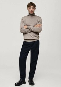 Beige polokrage tröja i ribbat material, kombinerad med mörkblå byxor och svarta snörskor, mot en enkel ljus bakgrund.