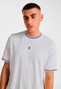 White short-sleeve t-shirt with navy-striped collar and sleeves; features a small black chevron logo on the chest. Made of lightweight fabric.