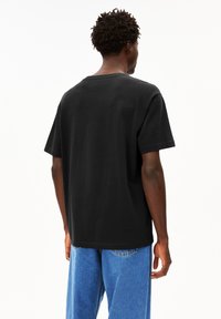 Schwarzes Baumwoll-T-Shirt mit lockerem Schnitt, kurzen Ärmeln und Rundhalsausschnitt, von hinten gezeigt. Getragen mit hellblauen Jeans.