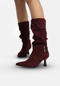 Bottines en daim bordeaux au design décontracté, bout pointu, accents décoratifs cousus, et petit talon avec détail de boucle en métal.