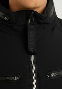 Nahaufnahme eines schwarzen Jacketts mit zentralem silbernem Reißverschluss und Zuglasche sowie zwei horizontalen Reißverschlusstaschen in Brusthöhe vor hellem Hintergrund.