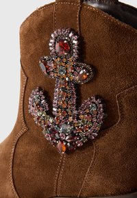 Brown suede boot with an ornate anchor-shaped embellishment made of multicolored red, orange, pink, and gray gemstones.